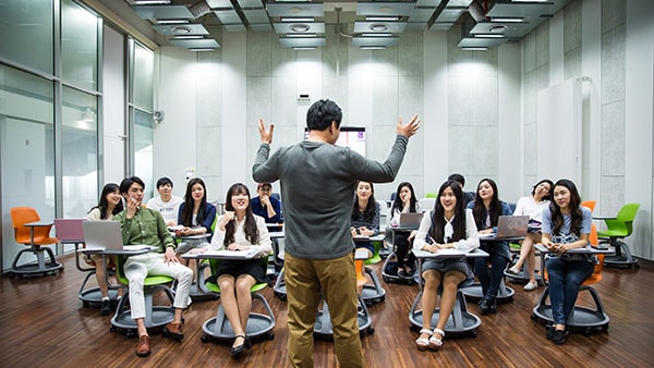 A teacher with arms raised presents to a group of students seated at desks in a classroom.