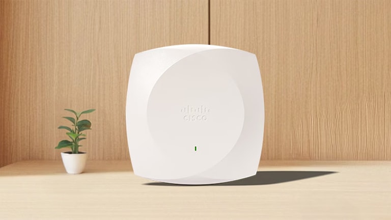 White Cisco wireless access point on a wooden desk next to a small potted plant.